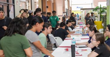 Decenas de personas siendo atendidas por personal reclutador de diversas empresas en el municipio de La Paz. El frente de la imagen destaca a tres jóvenes estudiantes recibiendo folletos informativos.