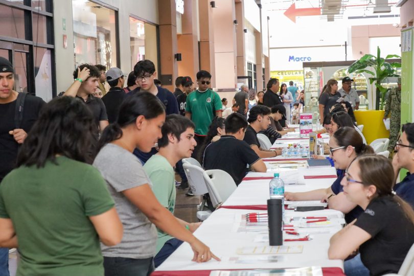 Decenas de personas siendo atendidas por personal reclutador de diversas empresas en el municipio de La Paz. El frente de la imagen destaca a tres jóvenes estudiantes recibiendo folletos informativos.